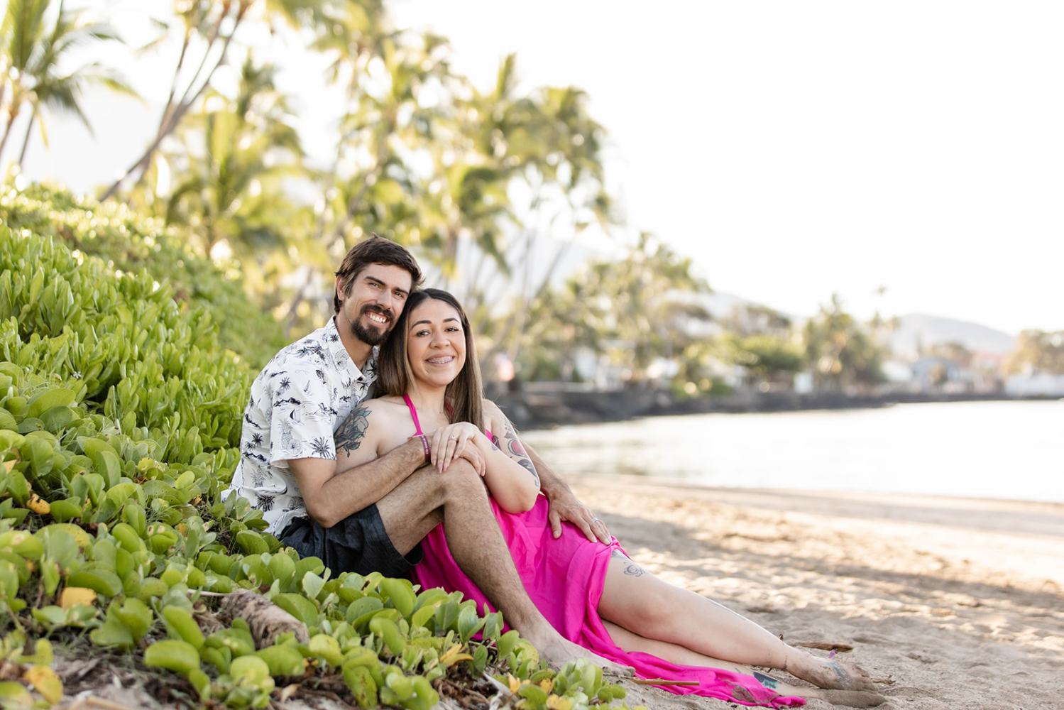 Couple sitting on a beach surrounded by greenery, smiling under a clear sky.