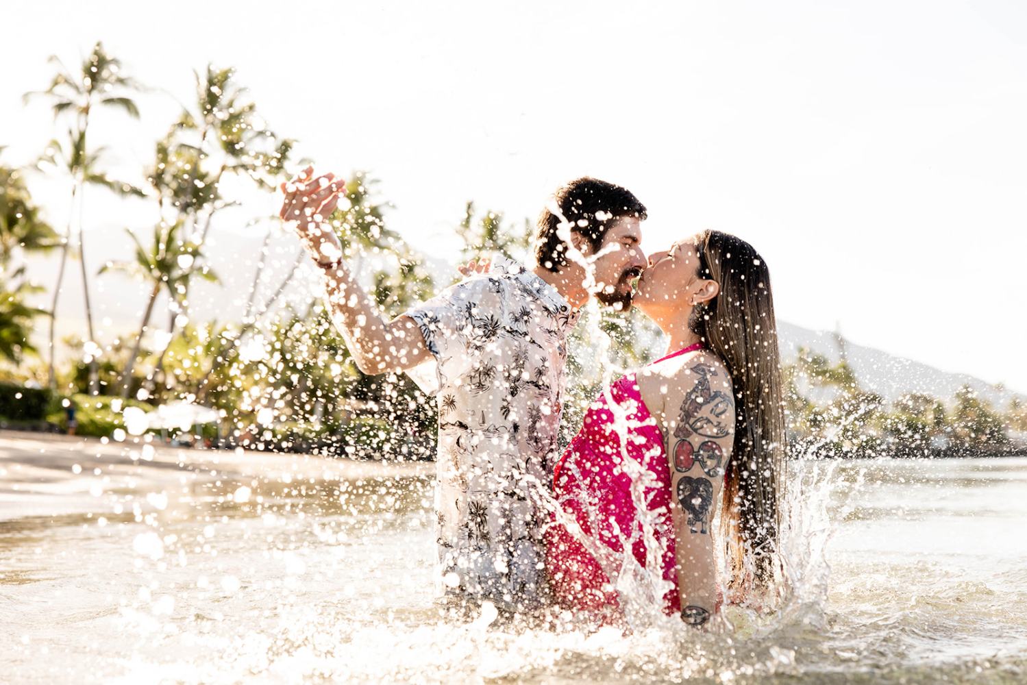 Couple kissing in ocean water splash, palm trees and mountains in background.
